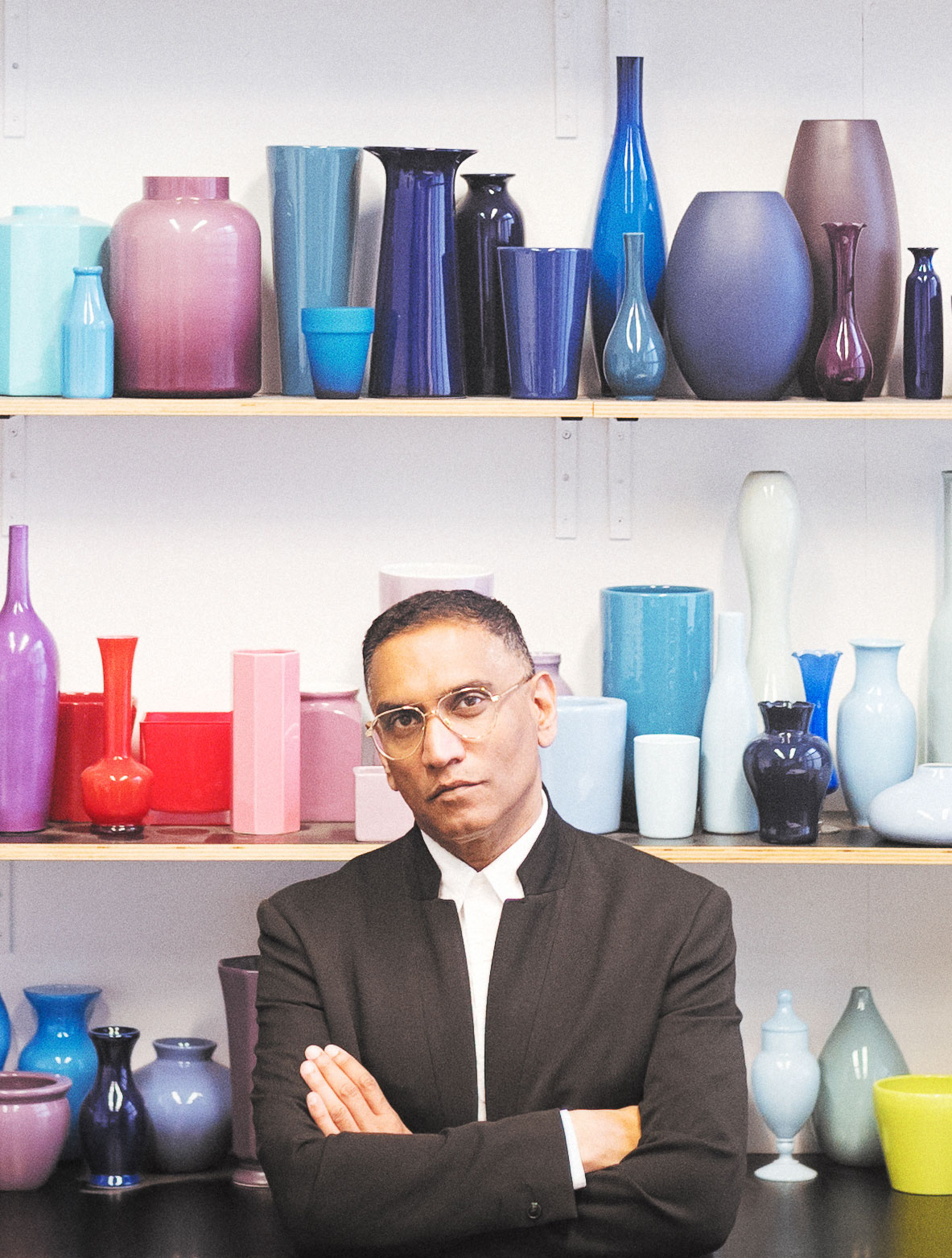 Portrait of artist David Sequiera standing in front of shelves lined up with colourful ceramics and glassware 